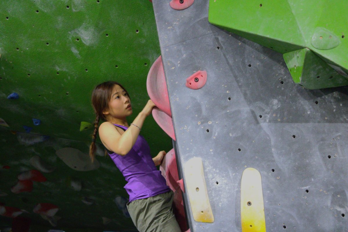 Raina Kim ('28) grips a red hold on a slanted wall, considering her next move during her ascent. The sport of bouldering involves more than physical strength to navigate sheer rock faces.